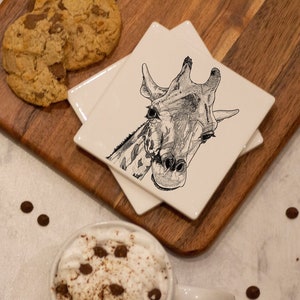 May include: A set of four white ceramic coasters featuring a black and white illustration of a giraffe's head on each coaster. The coasters are stacked on a wooden chopping board with a chocolate chip cookie on the board.