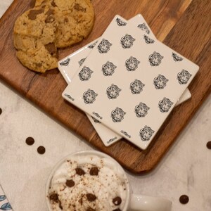 May include: A white gift box with a blue ribbon and a floral pattern, a mug of hot chocolate with whipped cream and chocolate sprinkles, two white ceramic coasters with a black tiger print, and two chocolate chip cookies on a wooden cutting board.