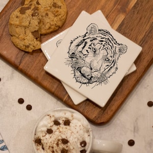 May include: A set of four white ceramic coasters with a black and white line drawing of a tiger's face on each coaster. The coasters are stacked on a wooden cutting board. There is a gift box wrapped in blue ribbon and a cup of coffee with whipped cream and chocolate sprinkles on a white surface.