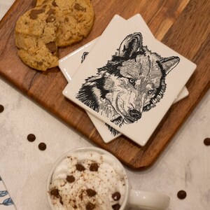 May include: A set of four white ceramic coasters with a black and white illustration of a wolf's head on each coaster. The coasters are stacked on a wooden cutting board with a chocolate chip cookie on the board. A cup of coffee with whipped cream and chocolate chips is on a white surface with chocolate chips scattered around.