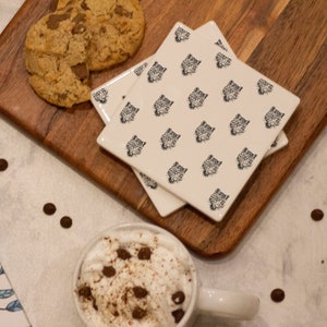 May include: A white gift box with a blue ribbon and a floral design, a mug of hot chocolate with whipped cream and chocolate sprinkles, two white ceramic coasters with a black leopard print design, and two chocolate chip cookies on a wooden chopping board.