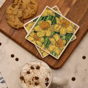 May include: A still-life arrangement featuring a mug of whipped cream topped with chocolate chips, a gift box tied with a blue ribbon, and two coasters with yellow daffodil designs. Chocolate chip cookies and pens are also present.