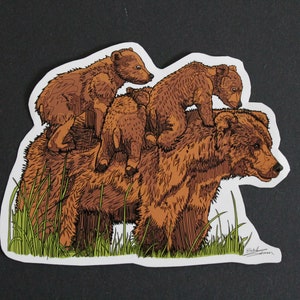 May include: A sticker featuring a brown bear with three cubs riding on its back. The illustration has a hand-drawn style with detailed linework and a touch of green grass at the bottom. The sticker is on a black background.