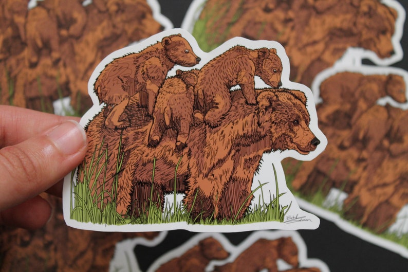 May include: A sticker featuring a brown bear with three cubs riding on its back. The illustration has a detailed, hand-drawn style with green grass at the bottom. The sticker is on a white background.