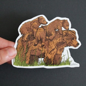 May include: A sticker featuring a brown bear with three cubs riding on its back. The illustration has a hand-drawn style with detailed fur and green grass at the bottom. The sticker is held against a dark background.