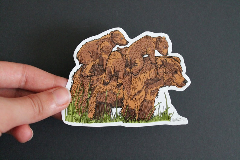 May include: A sticker featuring a brown bear with three cubs riding on its back. The illustration has a hand-drawn style with detailed fur and green grass at the bottom. The sticker is held against a dark background.