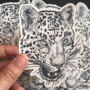 May include: A set of black and white line art stickers featuring a close-up of a leopard's face. The stickers are printed on white paper and have a glossy finish.