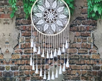 Peut inclure: Attrape-rêves en crochet blanc avec un motif circulaire et des glands blancs qui pendent du bas. L'attrape-rêves est suspendu à un mur de briques avec des vignes vertes.
