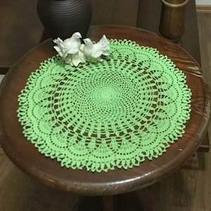 Op de afbeelding: Een groene gehaakte placemat ligt op een houten tafel met een vaas met roze lelies en twee witte keramische vogels.