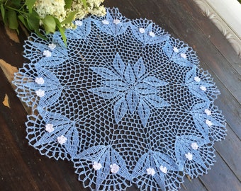 May include: A light blue, handmade crochet doily with a floral design. The doily features a central starburst pattern and is adorned with small, white floral accents around the edge. The intricate openwork design is set against a dark wooden surface.