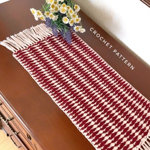 May include: A crocheted table runner with a diamond pattern in burgundy and white with a fringe border. The text "CROCHET PATTERN" is visible in the image.