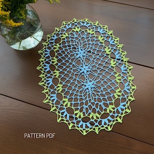 Op de afbeelding: Een blauwe en groene gehaakte ovale placemat met een delicaat bloemenpatroon. De placemat ligt op een houten oppervlak. PATTERN PDF