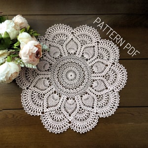 May include: A delicate pink crochet doily with a circular design. The doily is made with a fine thread and has a delicate, lacy appearance. The doily is laid out on a wooden surface. The text "PATTERN PDF" is visible in the upper right corner of the image.