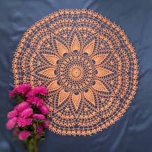 Op de afbeelding: Een delicaat perzikkleurig gehaakt doily met een cirkelvormig ontwerp en ingewikkelde bloemmotieven. Het doily ligt op een donkerblauwe stoffen achtergrond.