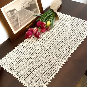 Könnte beinhalten: Ein weißer gehäkelter Tischläufer mit einem zarten Muster. Der Läufer liegt auf einem dunkelbraunen Holztisch mit einem Strauß roter und gelber Tulpen.