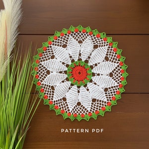Pode incluir: Um crochê branco com detalhes verdes e laranja. O crochê tem um design floral com uma flor central. O texto "PATTERN PDF" está impresso abaixo do crochê.