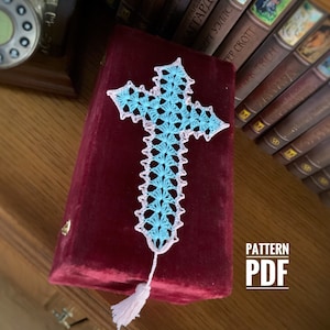 Puede incluir: Un marcapáginas de punto de cruz azul y rosa con un borla. El marcapáginas está sobre un libro de terciopelo rojo. El texto "PATTERN PDF" está impreso en el libro.