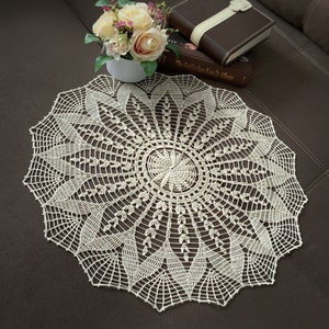 Può includere: Un tovaglietta all'uncinetto bianca con un delicato motivo floreale. La tovaglietta è circolare e ha un diametro di circa 30 cm.