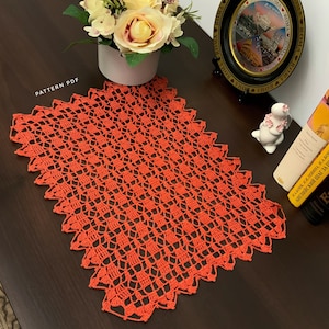May include: A bright orange crocheted table runner with a delicate pattern. The runner is laid out on a dark wood table.