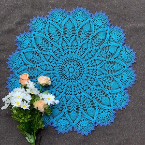 Peut inclure: Nappe ronde faite à la main, turquoise avec une bordure festonnée bleu foncé. Le motif de crochet complexe présente un design floral. Un petit bouquet de marguerites blanches et de roses pêche est posé sur le côté.