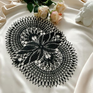 Puede incluir: Doily de crochet negro con un delicado patrón floral. El doily tiene forma ovalada y un diseño intrincado.
