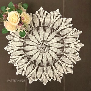 Peut inclure: Un napperon en crochet blanc avec un motif floral délicat. Le napperon est circulaire et a un bord festonné. Le napperon est posé à plat sur une surface brune.
