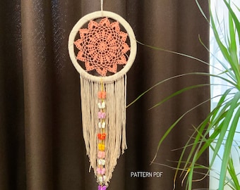 May include: A peach-colored crocheted dreamcatcher with white fringe and a string of colorful crocheted flowers hanging from the bottom. The dreamcatcher is hanging in front of a brown curtain. The text "PATTERN PDF" is visible at the bottom of the image.