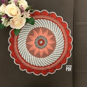 May include: A crocheted doily with a white centre and red, pink, and white concentric circles. The doily has a scalloped edge and is laid on a brown surface.