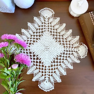May include: White crocheted doily with a delicate, intricate pattern. The doily is square-shaped with rounded corners and a central design.