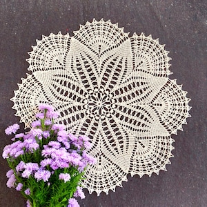 May include: A white crocheted doily with a delicate floral pattern. The doily is laid flat on a dark gray surface and a bouquet of purple flowers is in the foreground.