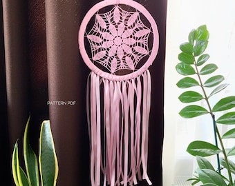 May include: A pink crocheted dreamcatcher with a large, intricate design and long, flowing ribbons. The dreamcatcher is hanging from a brown curtain.