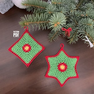May include: Two crocheted Christmas decorations, one a square with a red border and one a star with a red border. Both decorations are green with a yellow and red flower in the centre. The decorations are on a brown wooden surface with a green pine branch in the background.