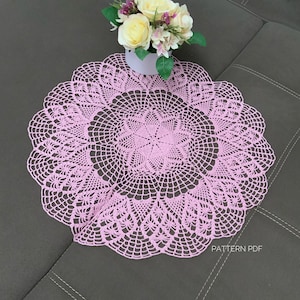Peut inclure: Un délicat napperon en crochet rose avec un motif floral. Le napperon est posé sur une surface grise avec un petit bouquet de fleurs blanches et roses dans un pot blanc.