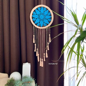 Pode incluir: Um apanhador de sonhos de crochê azul e branco com um aro de madeira e borlas bege penduradas nele. O apanhador de sonhos está pendurado na frente de uma cortina marrom. "PATTERN PDF" está escrito no canto inferior direito da imagem.
