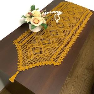 May include: A mustard yellow crocheted table runner with a diamond pattern and a tassel on one end. The runner is on a wooden table.