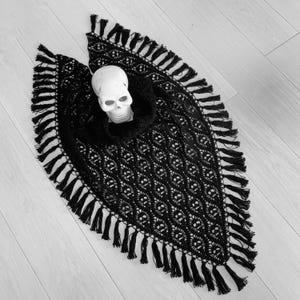 May include: A black, open-weave poncho with a skull-shaped design and fringe trim. A white skull is placed in the center of the poncho. The poncho is displayed on a light-colored wooden surface. The overall theme is gothic.