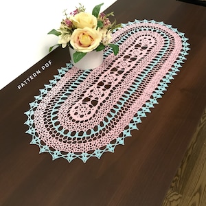 May include: A crocheted oval table runner in pink and blue with a delicate lace pattern. The runner is on a dark brown wooden table.