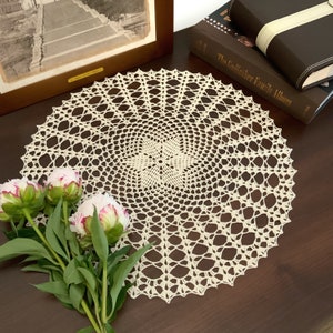May include: A delicate, off-white crocheted doily with a circular design. The doily is laid on a dark brown wooden surface with a bouquet of pink peonies in the foreground.