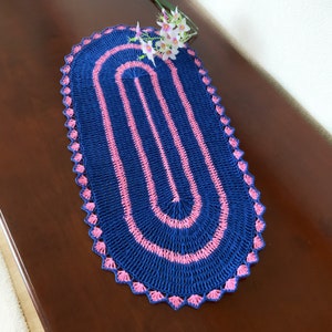 Peut inclure: Chemin de table ovale en crochet, bleu marine avec des détails roses. Le motif est composé d'ovales roses répétés, bordés d'une bordure festonnée. Un petit arrangement floral est posé sur la table.