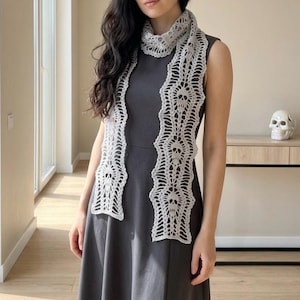 May include: A light gray, open-weave crochet scarf featuring a repeating skull pattern. The scarf is draped over a dark gray dress. The design is intricate and detailed, with a scalloped edge. The scarf appears to be handmade.