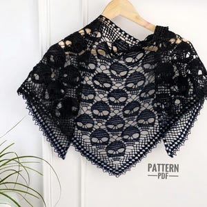 May include: A black triangular crochet shawl featuring a repeating skull motif throughout the fabric. The shawl is displayed on a wooden hanger and includes the text PATTERN PDF printed in the lower right corner of the image.