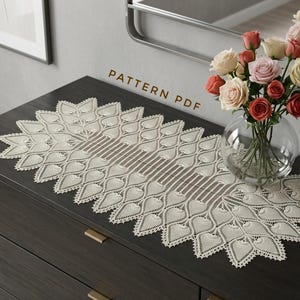Könnte beinhalten: Ein cremefarbener, ovaler Häkel-Tischläufer mit einem sich wiederholenden blattartigen Muster und komplizierten Durchbrucharbeiten. Der Text "PATTERN PDF" ist in Braun über dem Tischläufer auf einer dunklen Holzkommode abgedruckt.