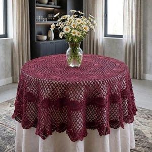May include: A round, burgundy crochet tablecloth with a detailed openwork design. The tablecloth is draped over a round table, with a vase of white daisies in the center. The table is set in a well-lit room with neutral-toned curtains.