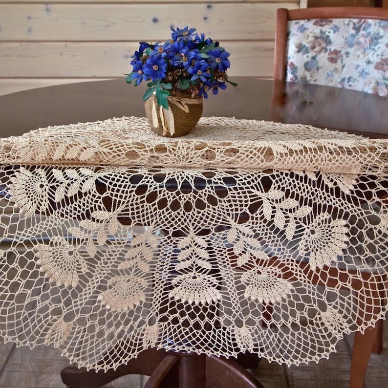 May include: A cream-coloured, round, crocheted tablecloth with a floral pattern, covering a table. A small woven basket with blue flowers is on top. A wooden chair with a floral back is in the background.