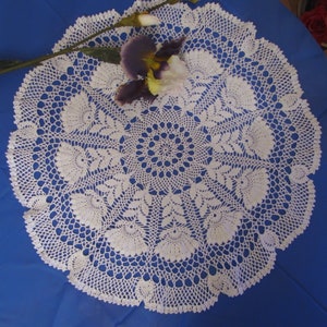 May include: A white, circular, crocheted doily with a floral design. The doily is placed on a blue surface and adorned with a purple and white flower and a stem. The intricate lace pattern is visible throughout the piece.