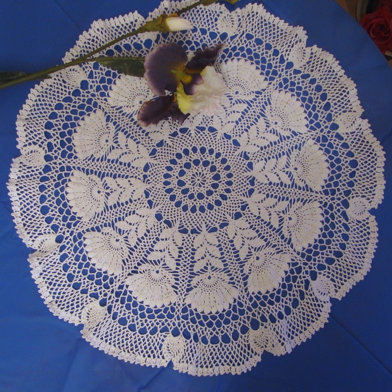 May include: A white, circular, crocheted doily with a floral design. The doily is placed on a blue surface and adorned with a purple and white flower and a stem. The intricate lace pattern is visible throughout the piece.