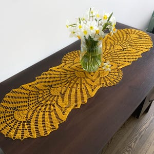 Puede incluir: Un camino de mesa de crochet amarillo con un patrón delicado e intrincado. El camino de mesa está colocado sobre una mesa de madera marrón oscuro con un ramo de flores blancas en un jarrón de vidrio.