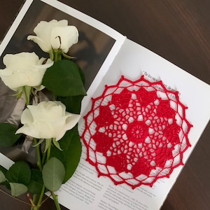 May include: A red crocheted doily with a delicate floral pattern, resting on a book with white roses in the background.