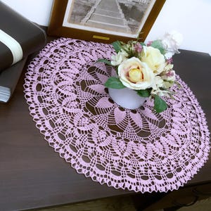 May include: A round, light pink crocheted doily with an intricate floral lace pattern. The doily is placed on a dark brown surface and is decorated with a small white vase of flowers.