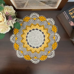 Op de afbeelding: Een wit en geel gehaakt tafellaken met een rond ontwerp. Het tafellaken heeft een centrale cirkel met een stervormig patroon en een gegolfde rand.